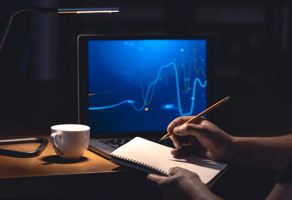 Investidor analisando gráficos financeiros em um notebook enquanto faz anotações em um caderno